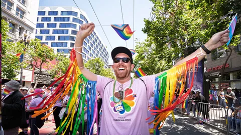A UCSF employee marching in the 2025 San Francisco Pride Parade, waving pride flags and wearing rainbow colored fringe on their arms