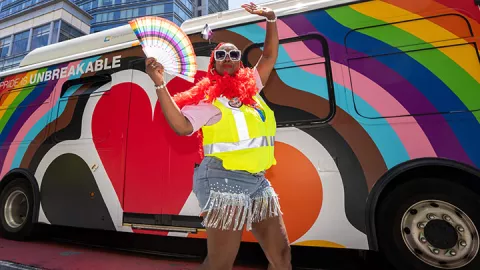 A UCSF employee wearing a yellow safety vest and waving a rainbow fan beside a UCSF shuttle decorated with the 2025 San Francisco Pride Parade colors and theme