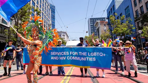 A Brazilian Samba dancer is waving a bisexual pride flag while marching in front of a contingent of UCSF community members holding an Alliance Health Project banner at the 2025 San Francisco Pride Parade